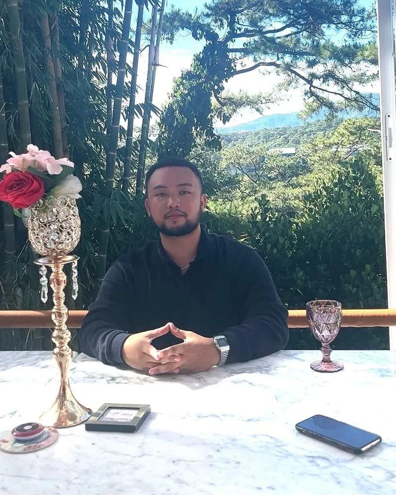 Christian Josef Aquino seated at a marble table, hands folded, with pine trees and hills behind him.